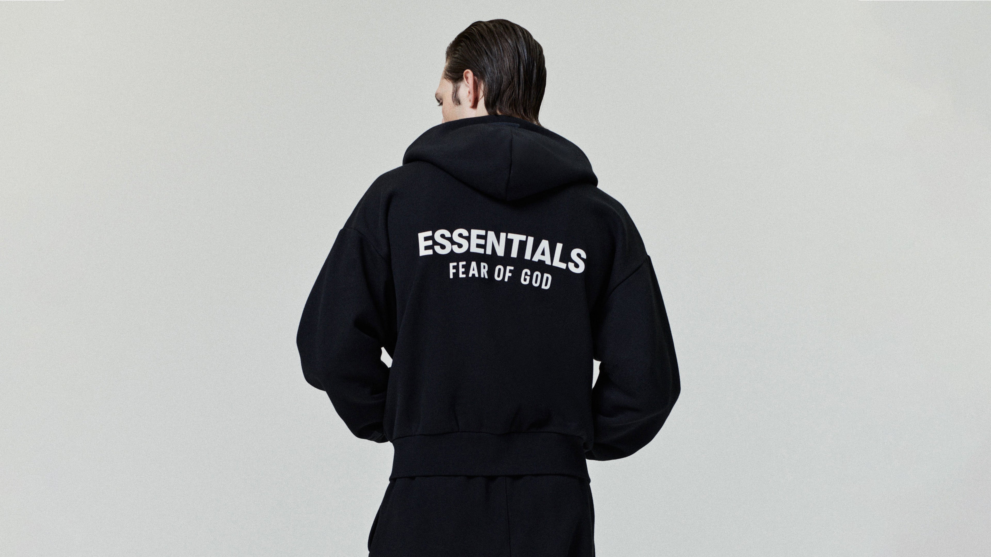 Person wearing a black hoodie with 'ESSENTIALS FEAR OF GOD' text on the back against a gray background