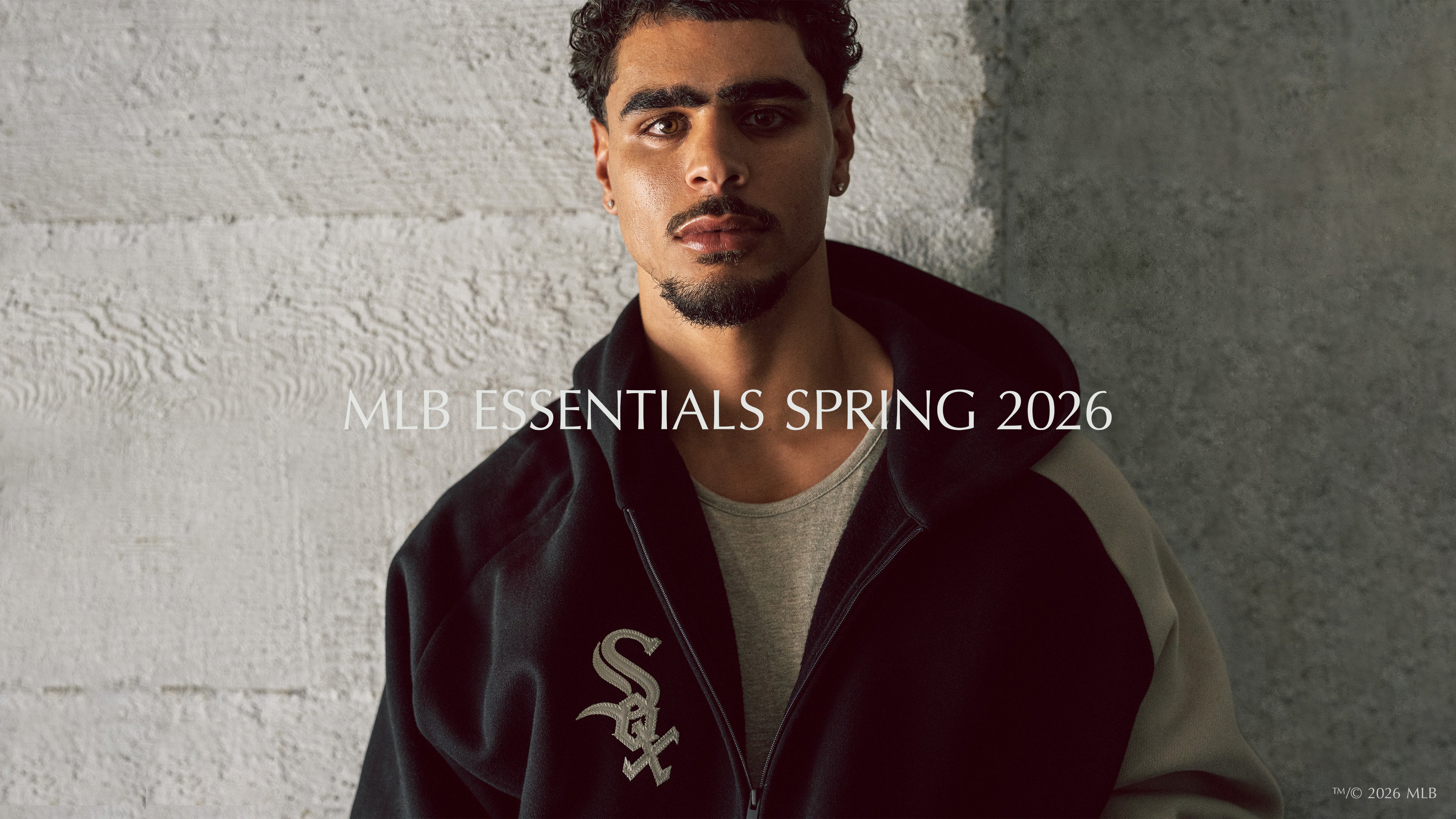 Portrait of a man with curly hair and a trimmed beard wearing a black and gray zip-up jacket with a white Chicago White Sox logo, standing against a rough concrete wall.