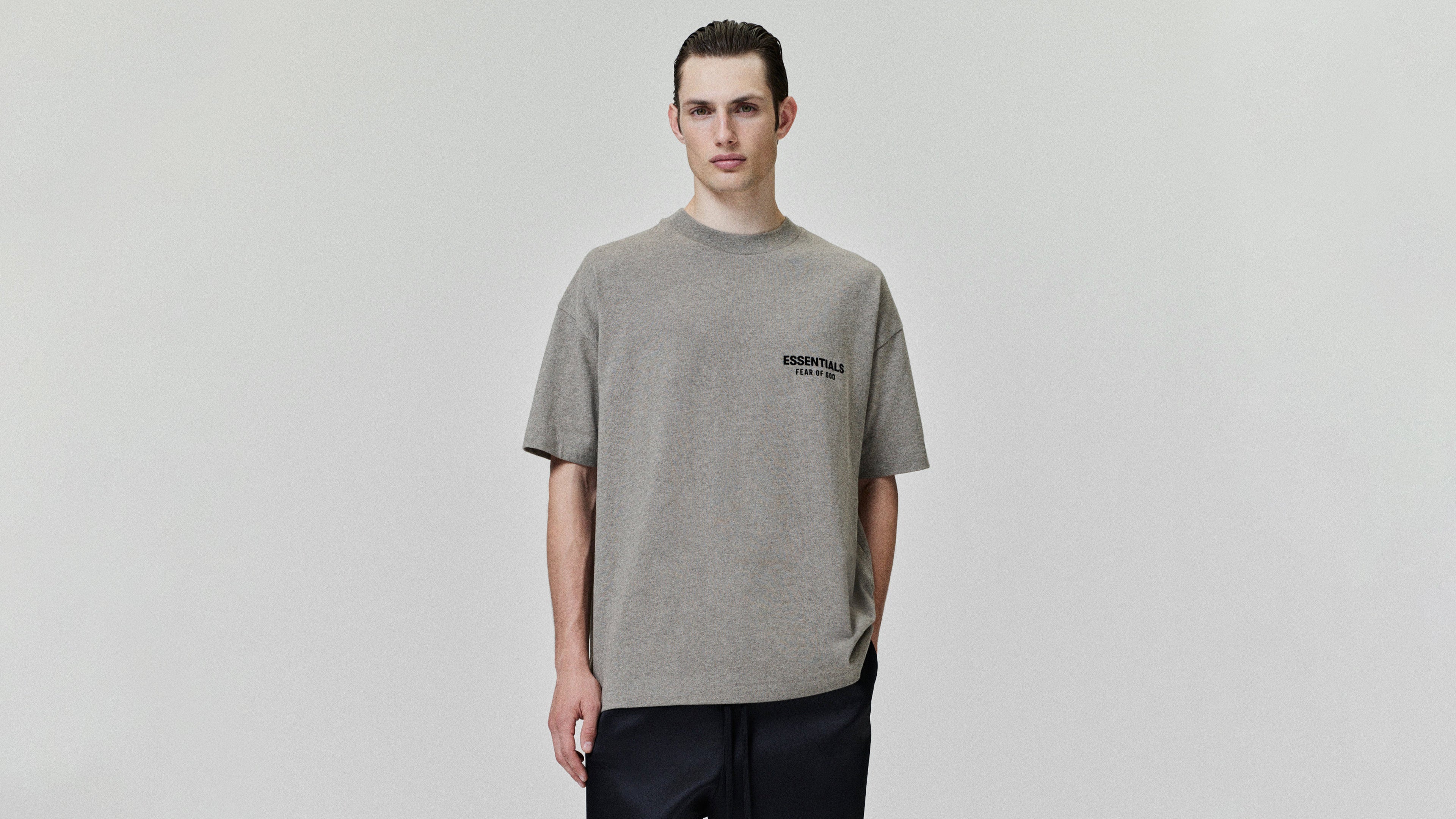 Person wearing a gray t-shirt with a logo reading "ESSENTIALS Fear of God" on a plain background